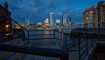 The photograph captures an urban architectural landscape of London’s Canary Wharf at dusk in summer, as seen from a riverside walkway on the north bank of the Thames. Taken in the evening, the image prominently features the illuminated Canary Wharf skyline framed by the surrounding railings and wooden deck, with distinctive buildings including One Canada Square and other modern high-rise towers. The River Thames stretches across the foreground, reflecting the city lights, while the pedestrian bridge to the right connects the riverside to the heart of the financial district. This architectural composition provides a clear view of London’s iconic business hub in midsummer, highlighting the interplay between the built environment and the waterway at a serene time of day.